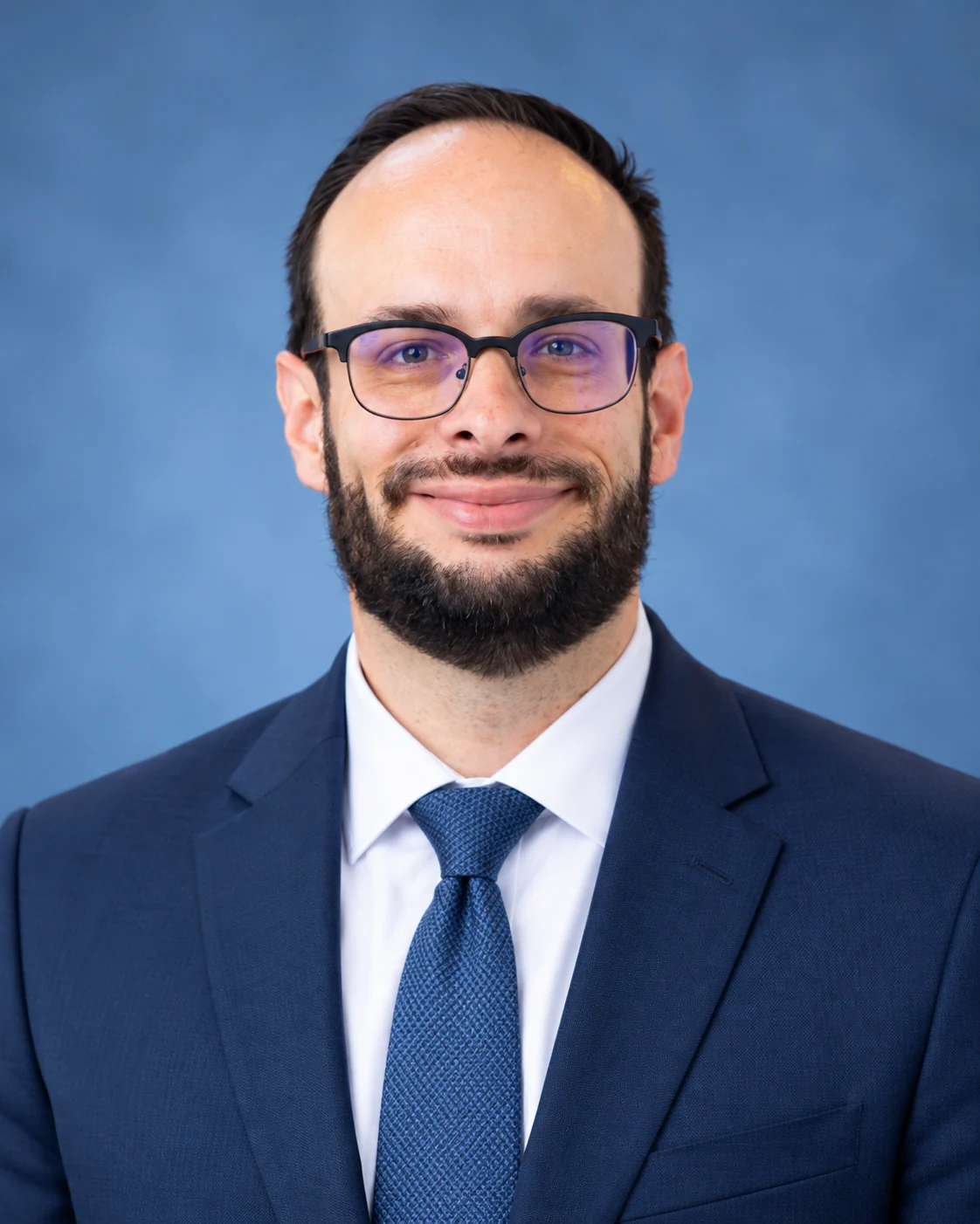 Donald Arsenault-Flachmeier, P.E., in a navy suit, blue tie, and glasses against a blue studio background.